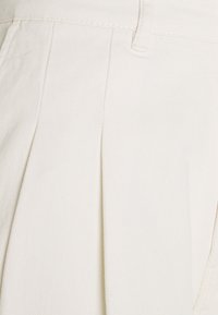 Pantalones plisados de color crema hechos de una tela texturizada, con bolsillos laterales y una cinturilla suave.
