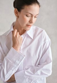 Biała koszula na guziki z klasycznym kołnierzykiem, długimi rękawami i gładką fakturą. Charakteryzuje się luźnym krojem i widocznymi guzikami na mankietach.