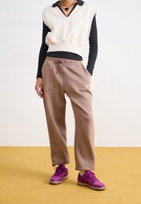 Person wearing beige sleeveless sweater over black long-sleeve top, brown Nike sweatpants, and purple Nike sneakers on yellow floor.