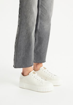 Sneaker bianche in pelle con superficie testurizzata e suola spessa, abbinate a jeans grigi a coste con orlo grezzo e dettagli di cucitura visibili.