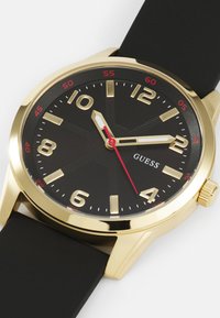 Black dial watch with a gold stainless steel case, prominent gold hour markers, red accents, and a black rubber strap.