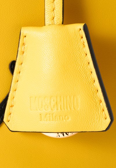 Étiquette en cuir jaune avec bords cousus, embossée avec "Moschino Milano" sur un fond en cuir jaune assorti.
