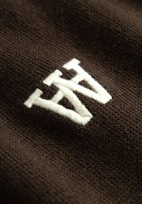 Brauner Stoff mit einer weichen Textur weist ein markantes, weiß sticktes Logo auf, welches ein stilisiertes "AW"-Design präsentiert.