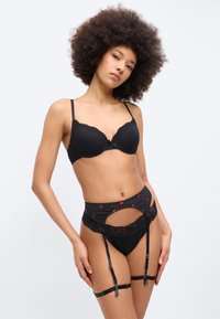 Woman with natural curly hair wearing black bra, garter belt with red floral details, black lace panties, and thigh straps, posing against plain background.