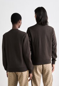 Brune genser med rund hals laget av bomullsblanding, med ribbestrikkede mansjetter og kant, sammen med mønstrede beige bukser.