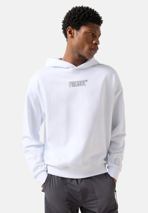 Junger Mann trägt einen weißen Hoodie mit dem Schriftzug "FIRE&ICE", die Hände in den Taschen, schaut seitlich vor einem einfarbigen hellen Hintergrund.