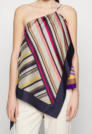 Frau trägt ein ein-ärmeliges, drapiertes Oberteil mit mehrfarbigen diagonalen Streifen und marineblauem Rand, kombiniert mit weißen Hosen.