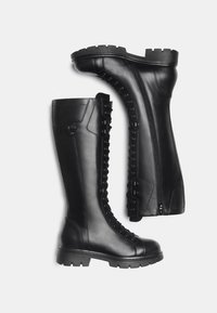 Des bottes en cuir noir arrivent au genou, avec une surface texturée, une fermeture éclair latérale et des lacets à l'avant. Semelle en caoutchouc épaisse avec grip à motifs.