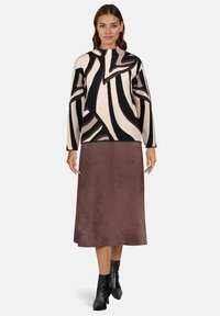 Vrouw draagt zwarte en beige abstracte print trui, bruine midirok en zwarte enkellaarzen, staande tegen een effen achtergrond.
