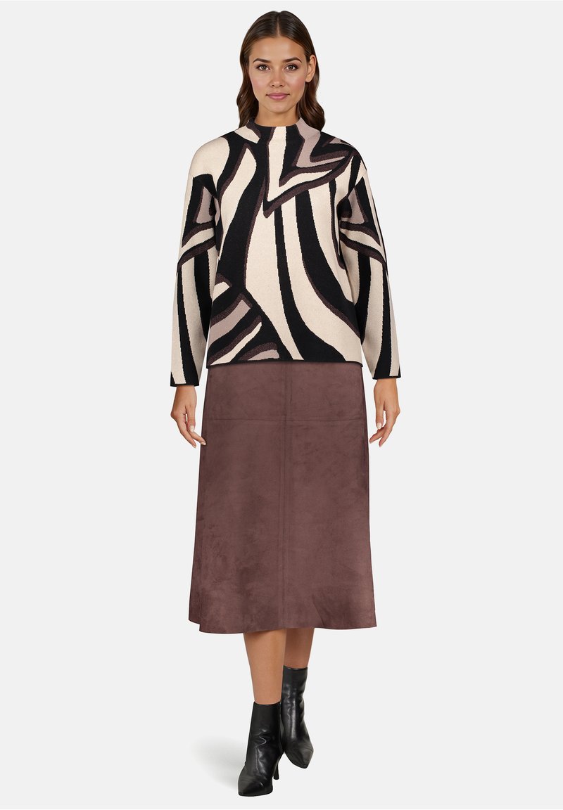 Vrouw draagt zwarte en beige abstracte print trui, bruine midirok en zwarte enkellaarzen, staande tegen een effen achtergrond.
