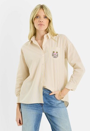 Mujer rubia con camisa de rayas verticales beige y blancas, con una pequeña herradura bordada y una flor en el bolsillo del pecho, y vaqueros azules.