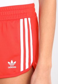 Red Adidas shorts with white three stripes on the side and the Adidas logo near the bottom hem, worn by a person with arm visible.