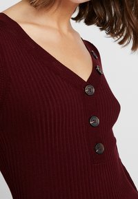 Maglione a coste bordeaux con scollo a V, caratterizzato da una serie di bottoni neri lungo un lato. La texture è morbida e aderente.