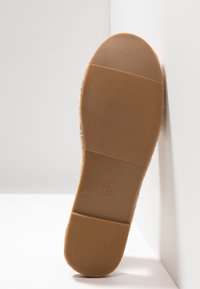 Semelle de chaussure en caoutchouc marron avec grip texturé, taille 4 indiquée, positionnée verticalement contre un mur d'angle blanc et un sol.