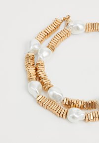 Necklace u zlatnom tonu s izmjeničnim pravokutnim i okruglim elementima, ukrašen s sjajnim bijelim biserima. Teksturalni zlatni dodaci dodaju detalj.