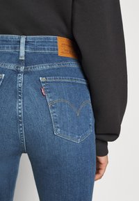 Sinised Levi's teksad kõrge vöökohtaga, tagakülgedel on taskud, millel on iseloomulik kõver õmblus, ja vöökohal pruuni nahast logo sildike.