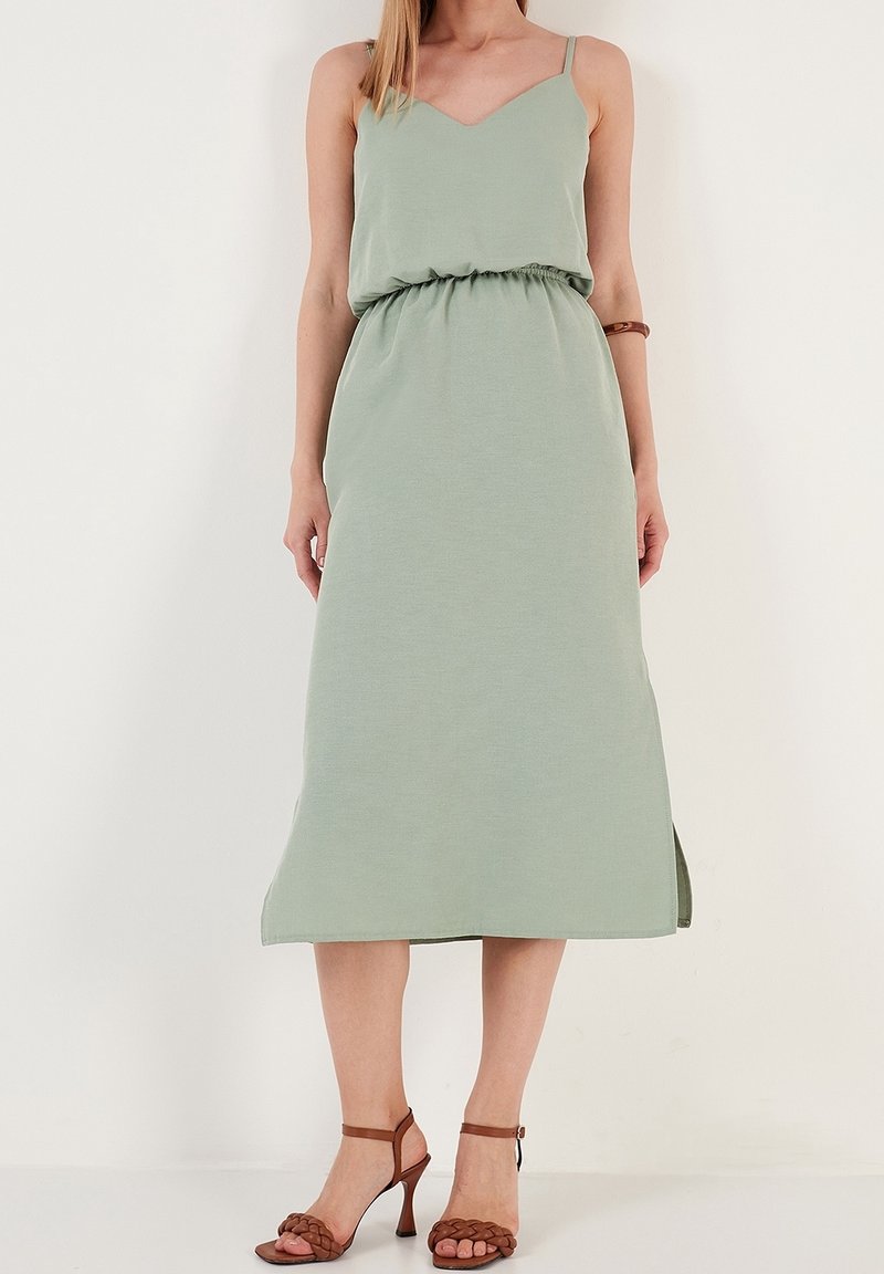 Femme portant une robe midi sans manches vert clair avec une taille cintrée et des sandales tressées marron à talons hauts, debout sur un fond blanc.