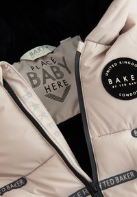 Vaaleanpunainen pehmeä vaate mustalla fleecevuorella. Siinä on "PLACE BABY HERE" -labeli ja pyöreä "BAKER by TED BAKER" -logo.