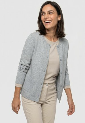 Vrouw met schouderlang donker haar draagt een lichtgrijze cardigan over een beige top en broek, glimlacht en kijkt opzij tegen een effen achtergrond.