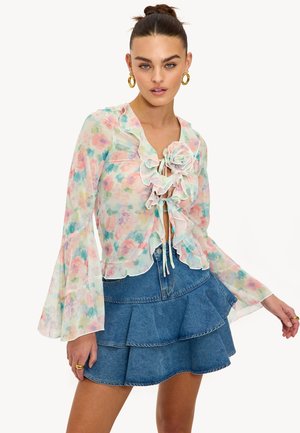Blouse fleurie à manches longues avec un col à volants et une fermeture à nouer, aux couleurs pastel. Associée à une mini-jupe en jean à volants.