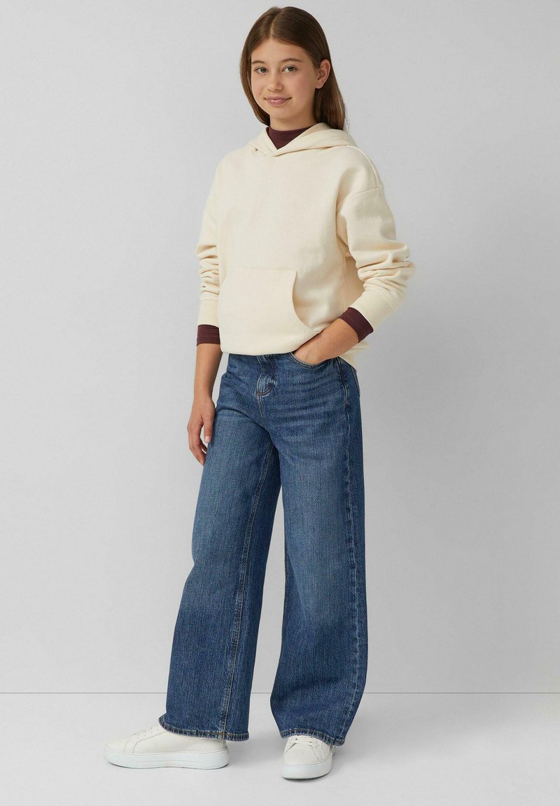 Crème hoodie met een voorzak en donkere manchetten, gecombineerd met een wijde blauwe jeans en witte sneakers op een lichtgrijze achtergrond.