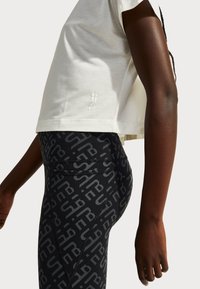 Close-up of person wearing white cropped top and black patterned leggings with abstract letters, standing sideways with arm relaxed.