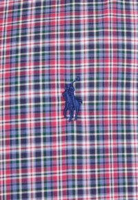 Rutigt tyg med röda, blå, gröna och vita rutor samt en marinblå broderad logotyp av en häst och ryttare på vänster bröst.