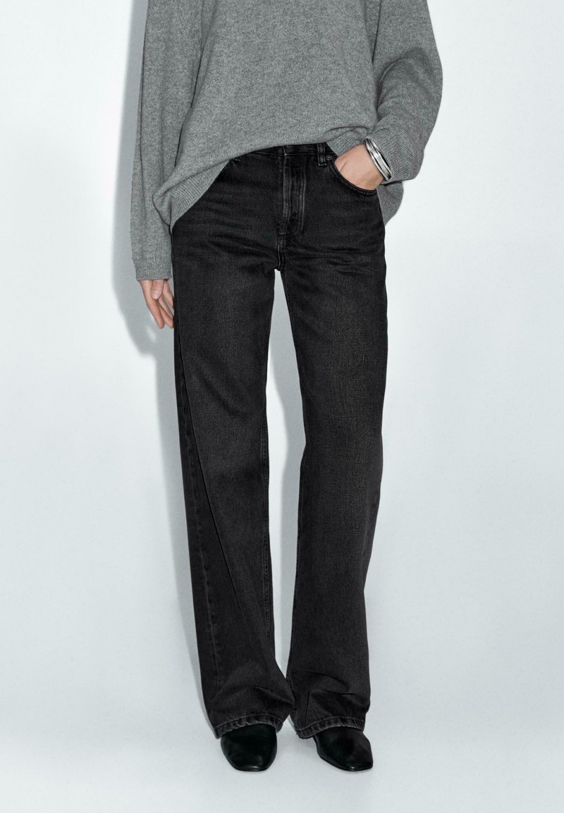 Jeans neri a gamba larga in denim, con vita alta e dettagli sbiaditi discreti. Abbinati a un maglione grigio e scarpe nere.