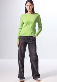 Maglione a coste verde lime con maniche lunghe, abbinato a jeans neri a vita alta con orlo sfilacciato e indossando sneaker metallizzate.