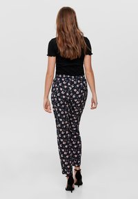 Haut noir texturé associé à un pantalon ajusté à motif floral en bleu marine foncé et rose. Modèle portant des talons noirs, vu de dos.