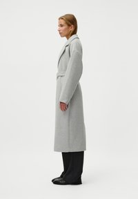 Cappotto grigio in lana con design avvolgente, maniche ampie e cintura. Il modello indossa pantaloni neri su misura e scarpe, visto di lato su uno sfondo bianco.