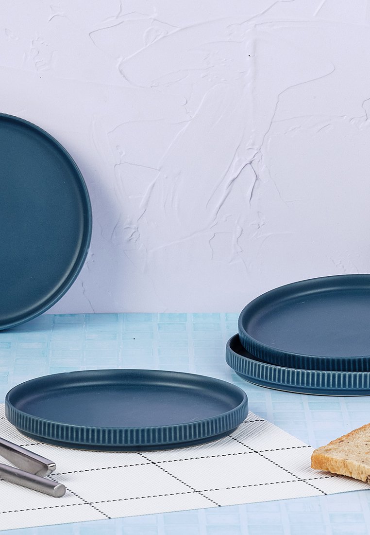 Ciemnoniebieskie ceramiczne talerze o gładkiej powierzchni i żebrowanych brzegach, ułożone z kawałkiem chleba i sztućcami na kratkowanej serwecie.