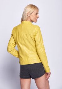 Gelbe Lederjacke mit tailliertem Design, hohem Kragen und genähten Details. Kombiniert mit dunkelgrauen Shorts.