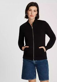 Czarna bluza z zamkiem na całej długości, z kołnierzem typu funnel, długimi rękawami i bocznymi kieszeniami, w połączeniu z dżinsowymi szortami. Gładka powierzchnia na całej długości.