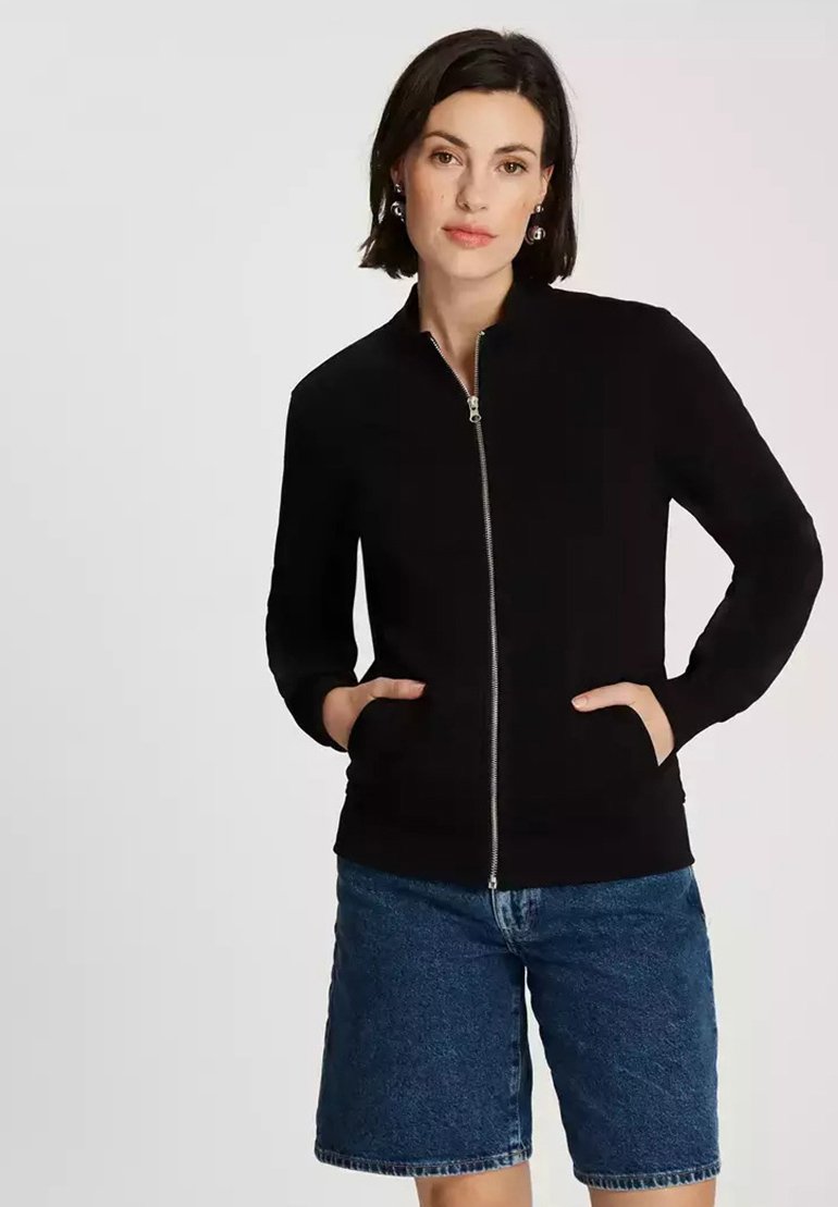 Czarna bluza z zamkiem na całej długości, z kołnierzem typu funnel, długimi rękawami i bocznymi kieszeniami, w połączeniu z dżinsowymi szortami. Gładka powierzchnia na całej długości.