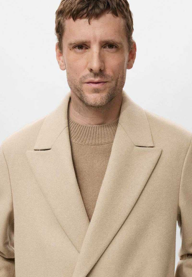 Beige wollen jas met ingekepte revers en enkele knoopsluiting, gelaagd over een tan geribde trui. Gladde textuur, slim fit, modern ontwerp.