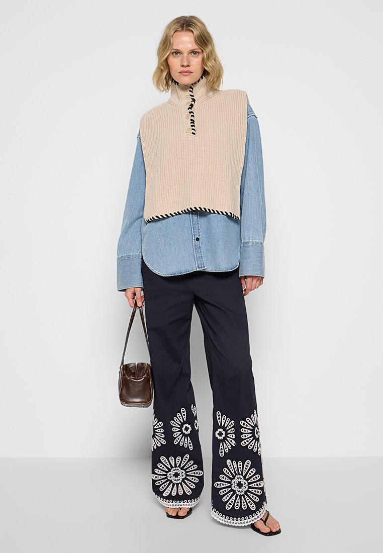 Beige gestrickter Poncho mit hohem Kragen, getragen über einem hellblauen Jeanshemd, kombiniert mit dunklen Hosen mit weißer Blumenstickerei.