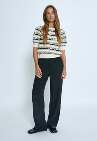 Gestrickter Pullover mit wellenförmigen schwarzen und cremefarbenen Streifen, mit Puffärmeln, kombiniert mit hoch taillierten schwarzen Anzughosen und glänzenden schwarzen Schuhen.