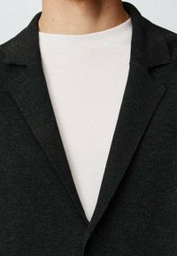 Blazer nero con rever a punta, tessuto texture e chiusura a un bottone, indossato sopra una maglietta a girocollo di colore chiaro.