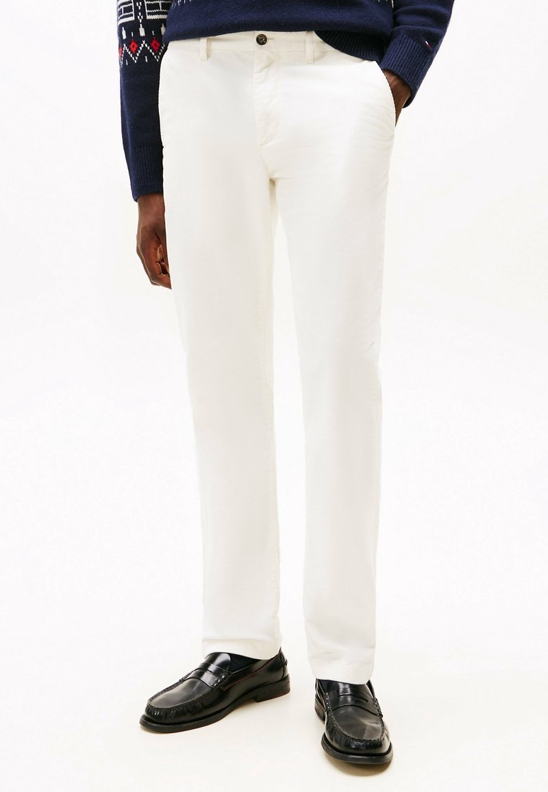 Homme portant un pantalon blanc droit, des mocassins en cuir noir et un pull bleu marine avec des manches à motifs, debout les mains dans les poches.