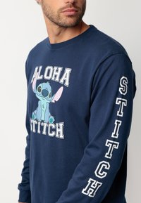 Camisa de manga larga color azul marino con un gráfico de Stitch, con el texto "ALOHA" y "STITCH" en blanco. Tejido de algodón suave, corte relajado.