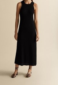 Robe midi noire à côtes avec une silhouette ajustée et sans manches. Assortie à des sandales noires à lanières. Texture lisse, élégance simple.