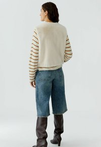 Nepbonten gilet in roomkleur over een beige gestreepte longsleeve, gecombineerd met lichtblauwe denim culottes en bruin leren laarzen tot de knie met hak.