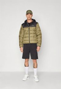 Oliven grønn vattert jakke med svarte skuldre, glidelås og hette. Brukt over svarte shorts med hvite detaljer og hvite sokker, stående mot en ensfarget bakgrunn.