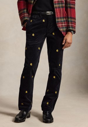 Homme portant un pantalon en velours côtelé noir avec de petits logos jaunes brodés, associé à une veste à carreaux rouge et des mocassins en cuir noirs.
