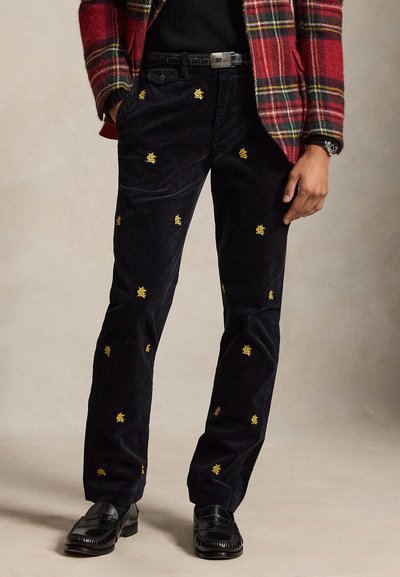 Άνδρας που φοράει μαύρο παντελόνι από κοτλέ με μικρά κίτρινα κεντημένα λογότυπα, συνδυασμένο με κόκκινο καρό σακάκι και μαύρα δερμάτινα μοκασίνια.
