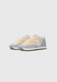 Par nogu bež i sivoj Saucony Jazz tenisicama sa detaljima od gamoša i bijelim srednjicama na jednostavnoj svijetlosivoj pozadini.