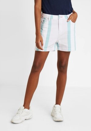 Person iført hvide shorts med lodrette lyseblå striber, hvide sneakers og en mørkeblå skjorte, stående med den ene hånd i lommen.