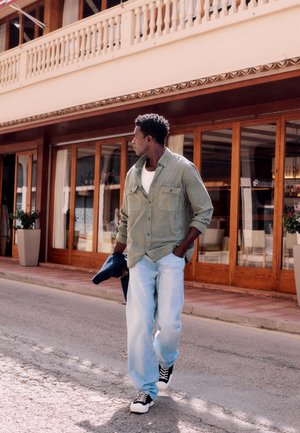 Mann in grünem Hemd und hellen Jeans geht die Straße entlang und hält eine marineblaue Jacke, im Hintergrund ein Holzladen und große Pflanzkübel.