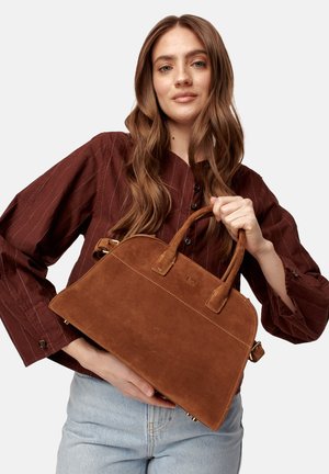 Giovane donna con lunghi capelli castani tiene una borsa in suede marrone, indossando una camicia a righe verticali rosso scuro e jeans azzurri su uno sfondo bianco.